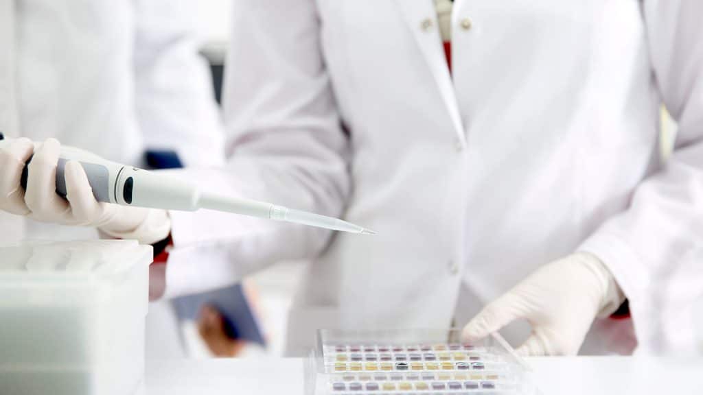 A person in a white lab coat holds a tester with a series of small test tubes sitting on a white counter in front of them.