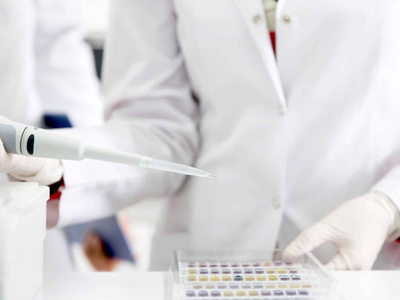 A person in a white lab coat holds a tester with a series of small test tubes sitting on a white counter in front of them.
