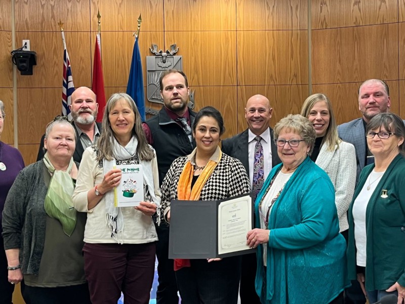 A group of older women stand with Fort St. John city council in a wood-paneled room. Everyone is looking at the camera