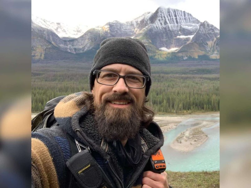 A man wearing a grey jacket and hat. He wears glasses and has a thick brown beard. He stands outside with water and mountains in the background.