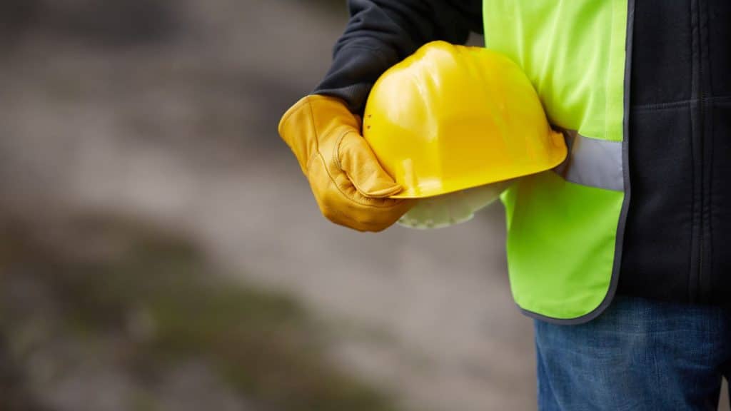 A person stands in a black sweater and yellow reflective vest. They hold a yellow safety helmet in their hand at their side.