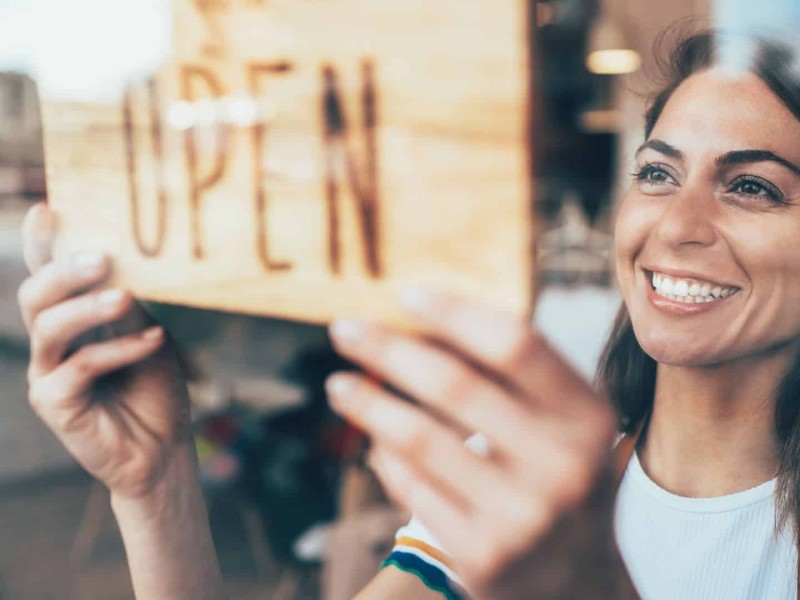 A smiling woman behind a pane of glass turns a small sign that says 'OPEN' on it.