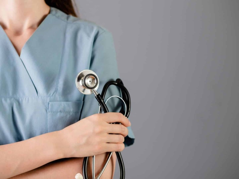 A woman stands in blue scrubs holding a stethoscope