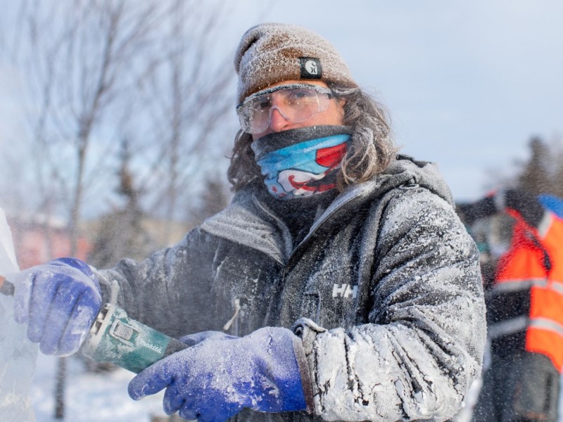 A man is winter gear holding a power tool to carve a block of ice.