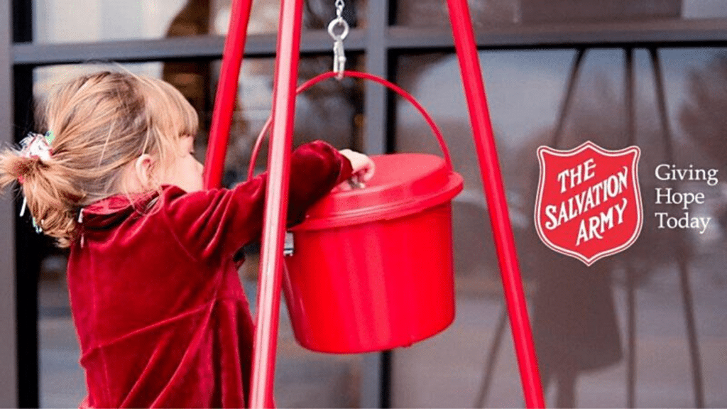 A small child in red putting money into a red Salvation Army kettle.