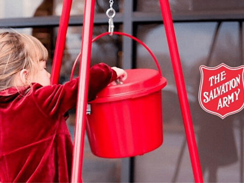 A small child in red putting money into a red Salvation Army kettle.