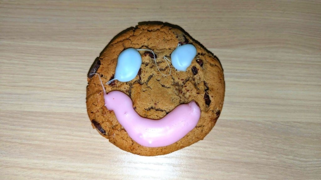 A chocolate chip cookie with a pink smile and blue eyes on a wooden desk.