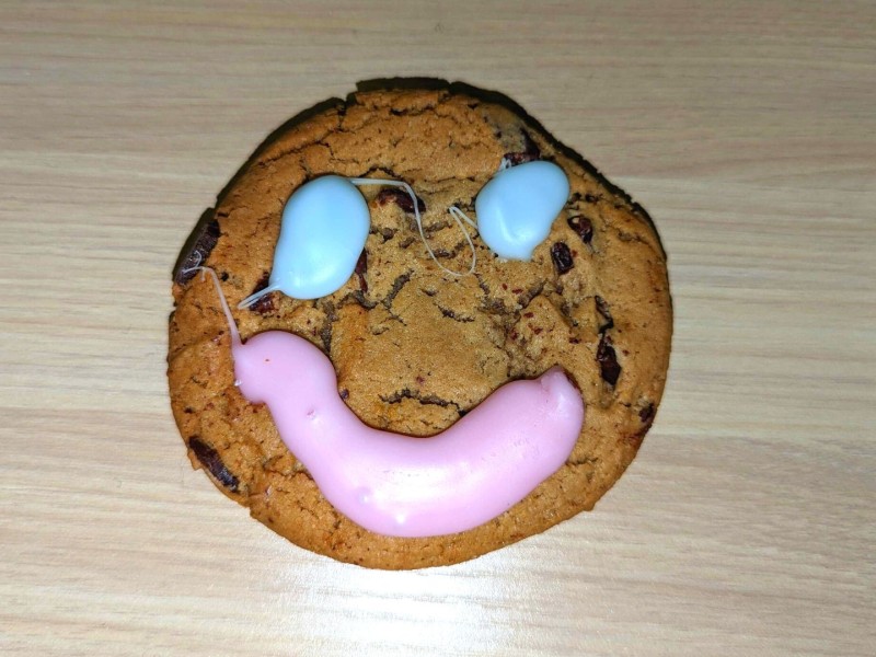 A chocolate chip cookie with a pink smile and blue eyes on a wooden desk.