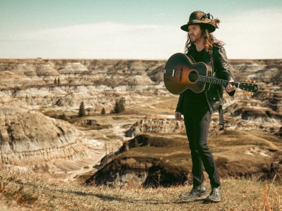 Kyle McKearney stands in front of a dessert wearing a cowboy hat and holding a guitar