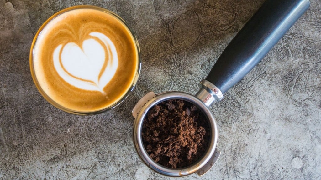 Coffee mug with a cream heart in it next to an espresso machine with coffee grounds in it on a grey background.