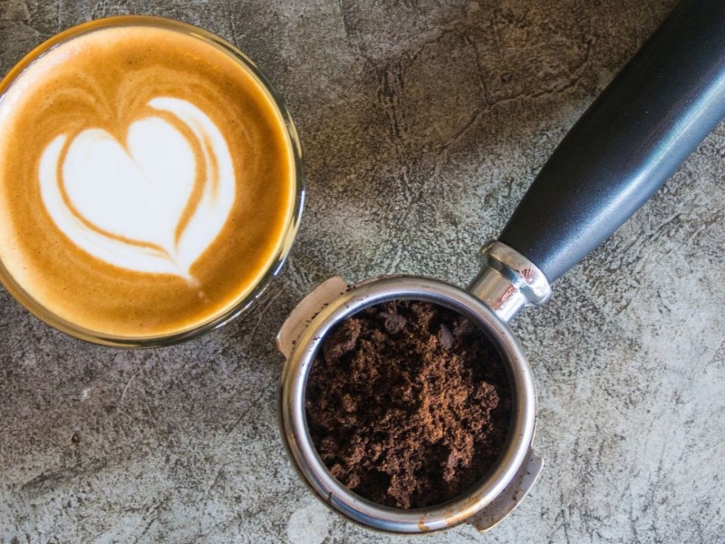 Coffee mug with a cream heart in it next to an espresso machine with coffee grounds in it on a grey background.