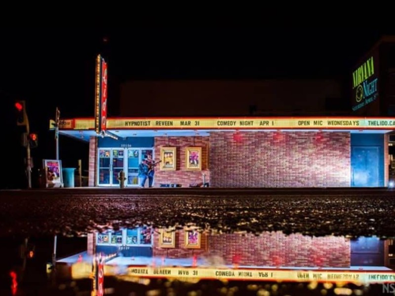A picture of Fort St. John's Lido Theatre front entrance lit up on a dark night, with a reflection of the brick building in a puddle.