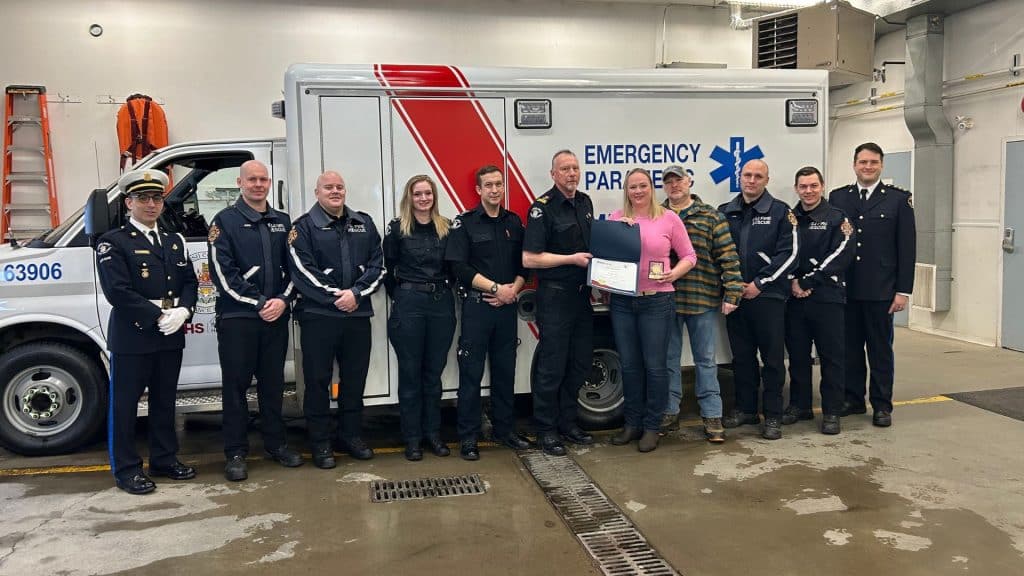 A group of people standing in front of an ambulance. Some paramedics, some firefighters, and a couple of civilians. The woman in pink is receiving an award.