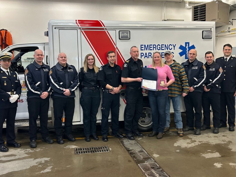 A group of people standing in front of an ambulance. Some paramedics, some firefighters, and a couple of civilians. The woman in pink is receiving an award.
