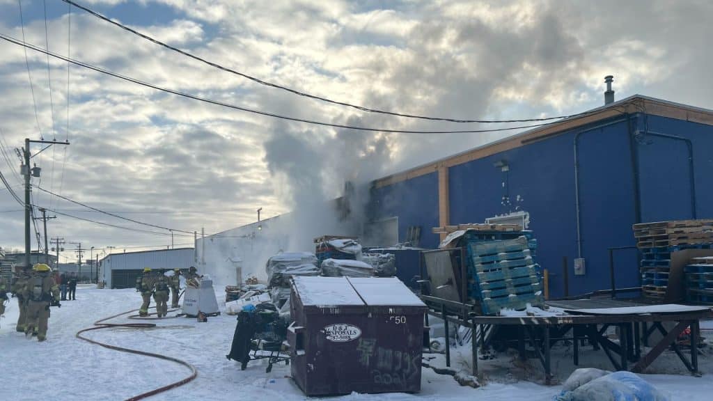 Firefighters standing outside of a blue building with smoke coming out the back of it.