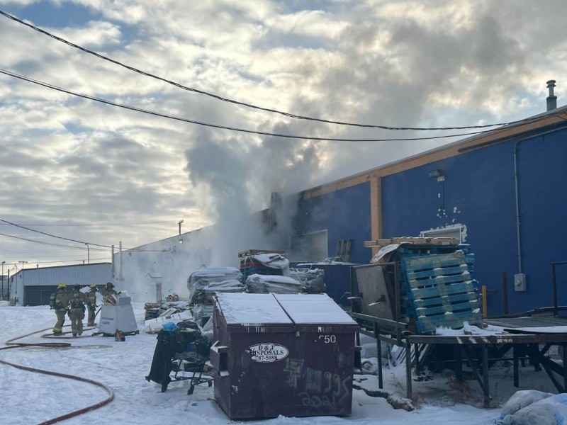 Firefighters standing outside of a blue building with smoke coming out the back of it.