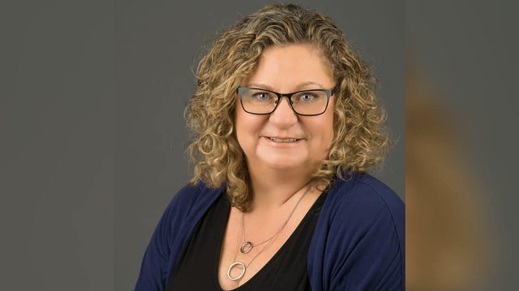 A headshot of a woman with blonde hair and glasses on a grey background.
