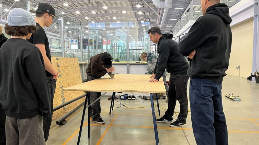 Students gathered around a sheet of wood. Two adults supervise as one of the students cuts the wood with a power tool.