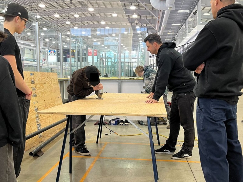 Students gathered around a sheet of wood. Two adults supervise as one of the students cuts the wood with a power tool.