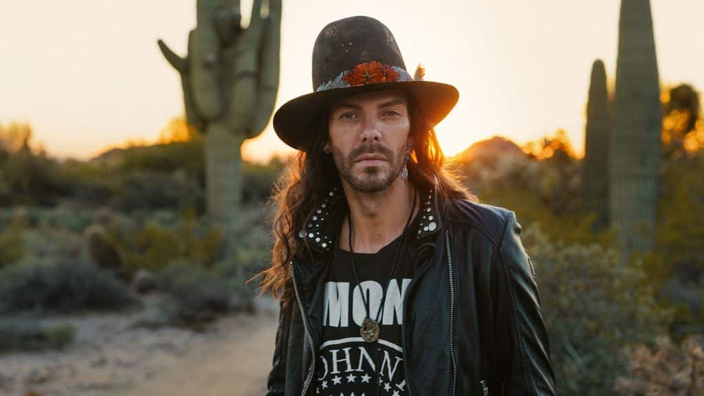 Photo of a man in a hat with facial hair standing in a desert with the sun behind him.