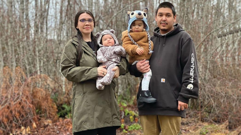 A young family with two children stand in the woods. Each parent holds one of the children.