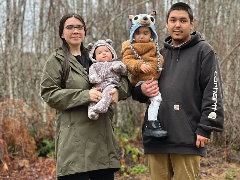 A young family with two children stand in the woods. Each parent holds one of the children.