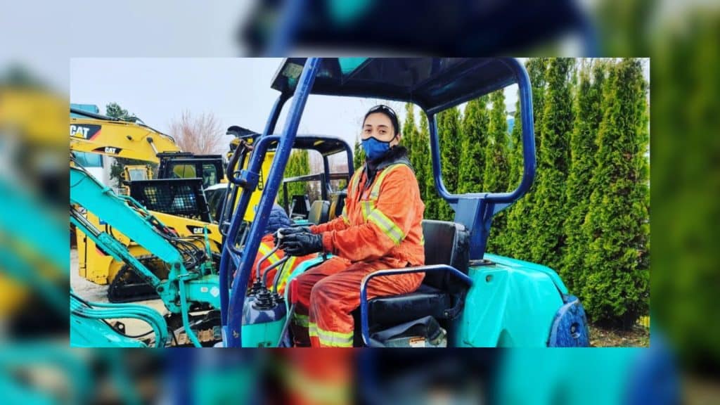 A woman in an orange and yellow safety jumpsuit, sits in a blue heavy machine.