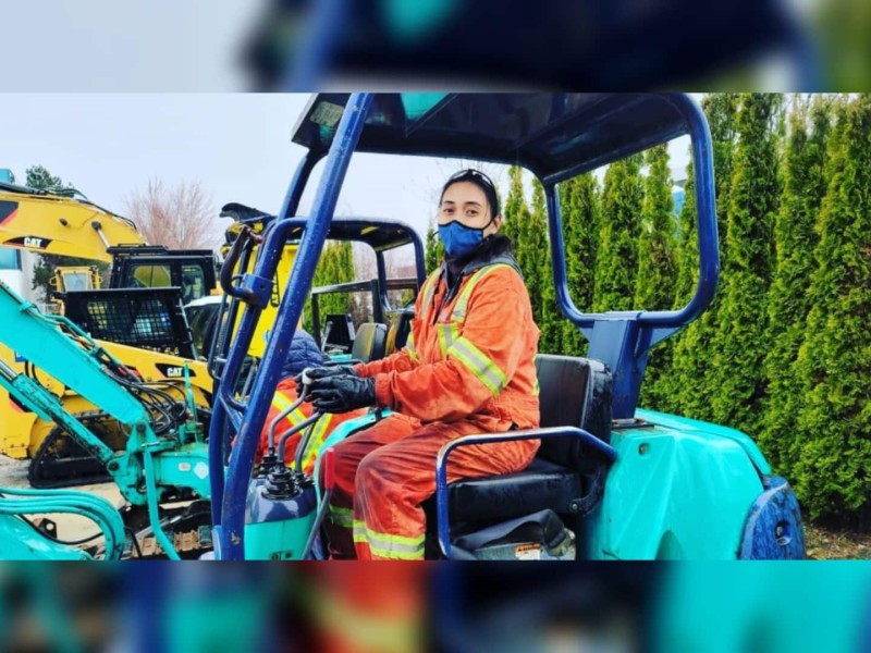 A woman in an orange and yellow safety jumpsuit, sits in a blue heavy machine.