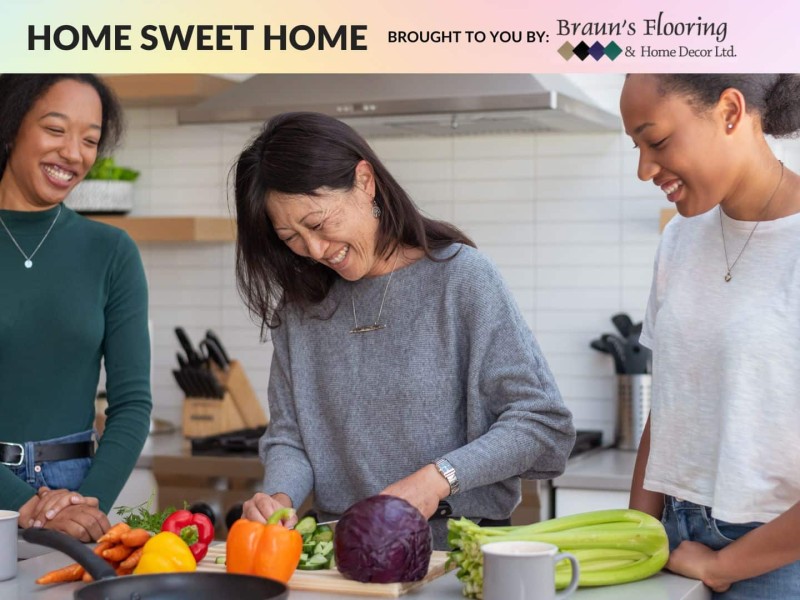 Three people standing in kitchen. Person on left smiling, person in middle cutting vegetables, person on right smiling
