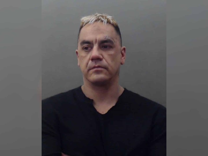 A blonde haired man in a black shirt on a grey background.