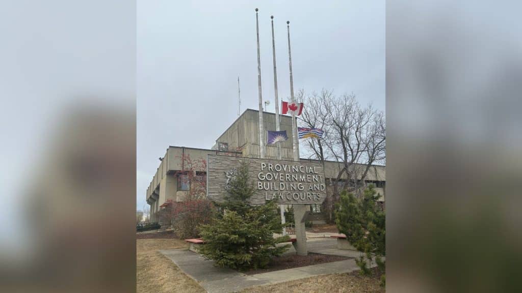 The Provincial Government Building and Law Courts sign in front of the grey brick building with the Fort St. John, Canada and B.C. flags at half mast.