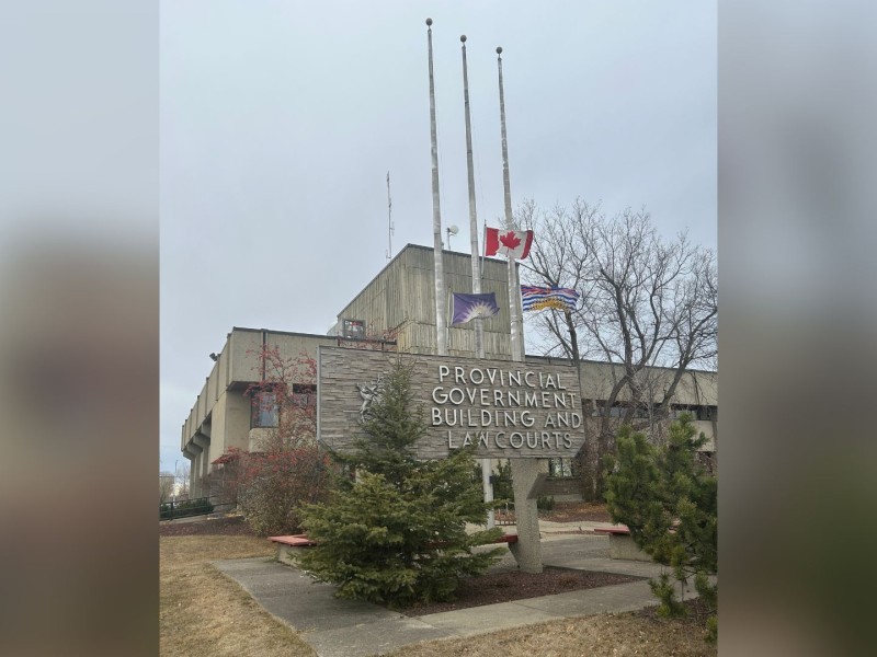 The Provincial Government Building and Law Courts sign in front of the grey brick building with the Fort St. John, Canada and B.C. flags at half mast.