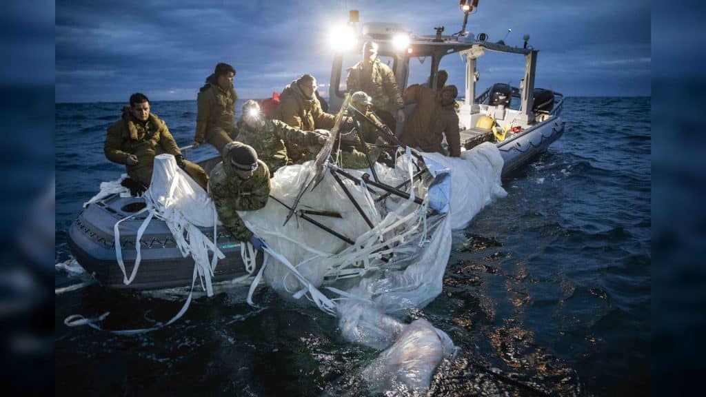 A group of soldiers in a small boat pull a large, white and dark metal apparatus out of the water.