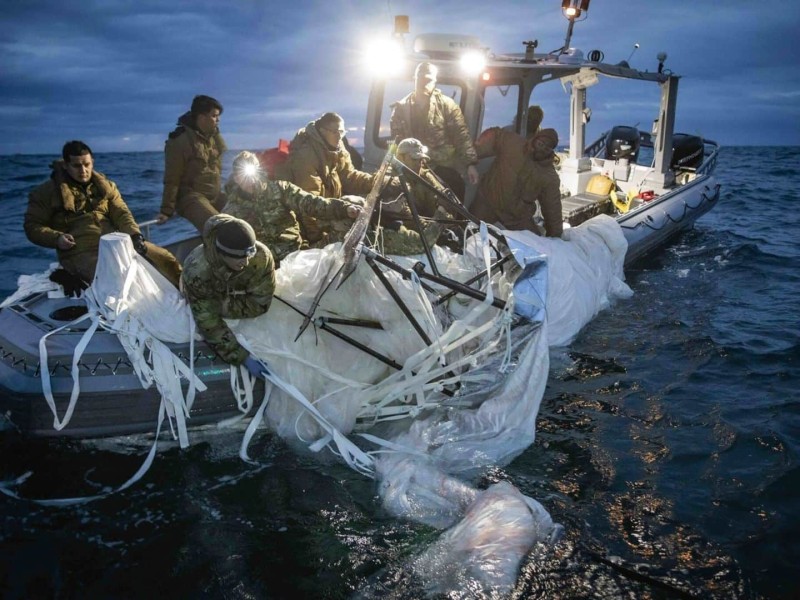 A group of soldiers in a small boat pull a large, white and dark metal apparatus out of the water.