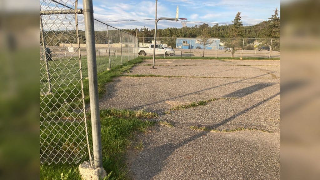 An old, asphalt basketball court surrounded by a chain-link fence and gross.