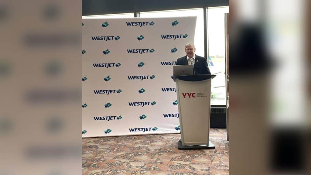 An old man in a suit stands behind a podium that reads 'YYC Calgary Airport Authority' in front of a WestJet background.