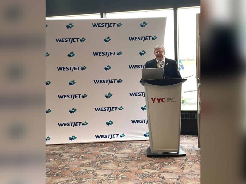 An old man in a suit stands behind a podium that reads 'YYC Calgary Airport Authority' in front of a WestJet background.