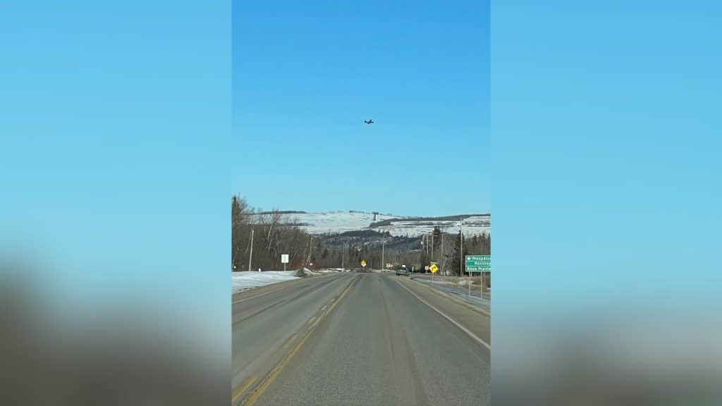 A plane flying  over some hills and mountains taken from inside a vehicle on the highway coming up to a sign.