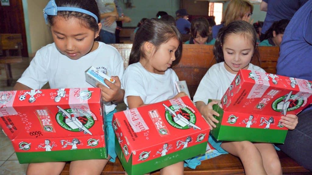 Three young children sit, opening red and green Christmas themed boxes.