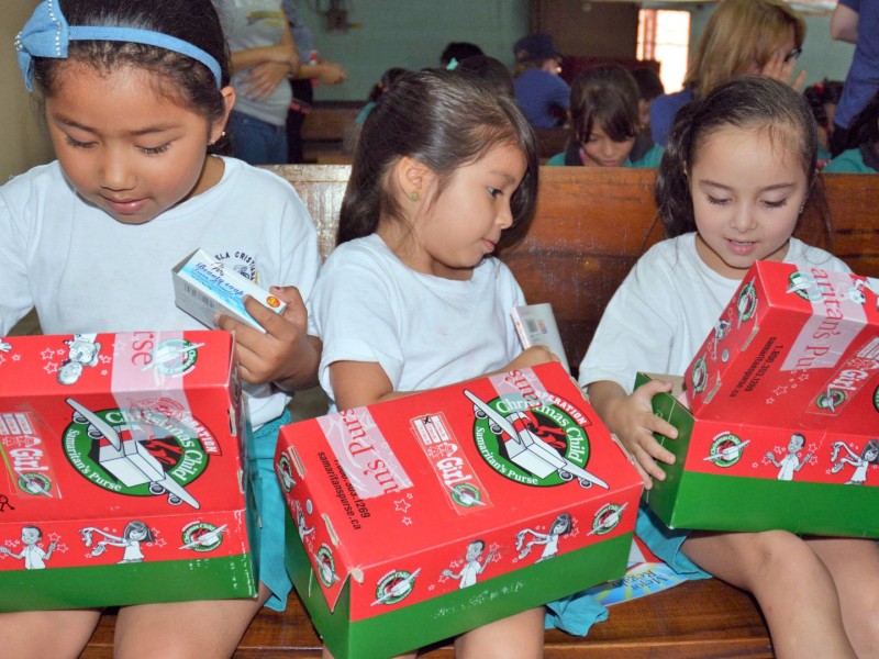 Three young children sit, opening red and green Christmas themed boxes.