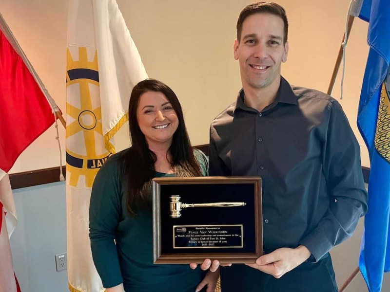 A man in a grey button-up and a woman in a blue shirt stand in frame in front of three flags. They hold an award that has a golden gavel on it.