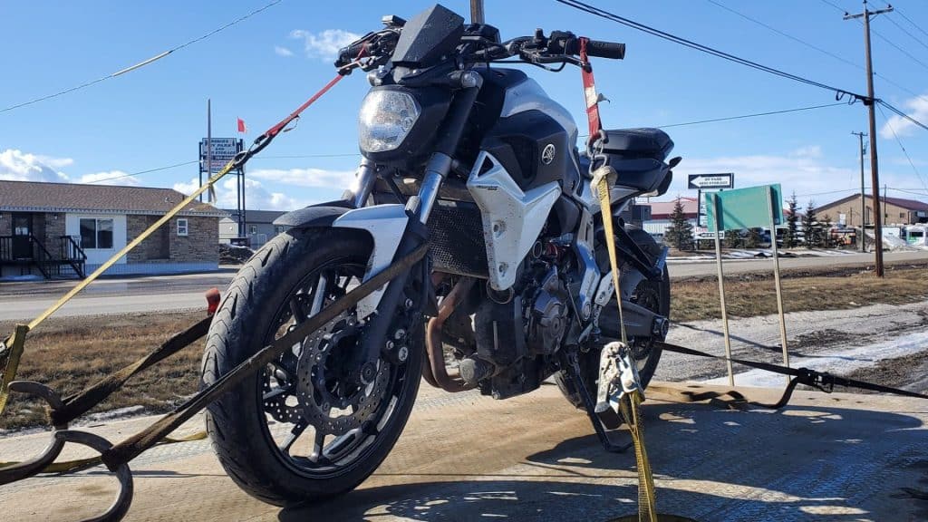 A motorcycle strapped to a tow truck bed on the roadside with buildings and signs in the background.