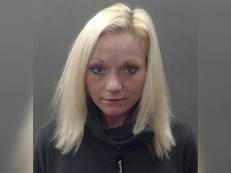 A blonde woman in a black sweater in front of a grey background.