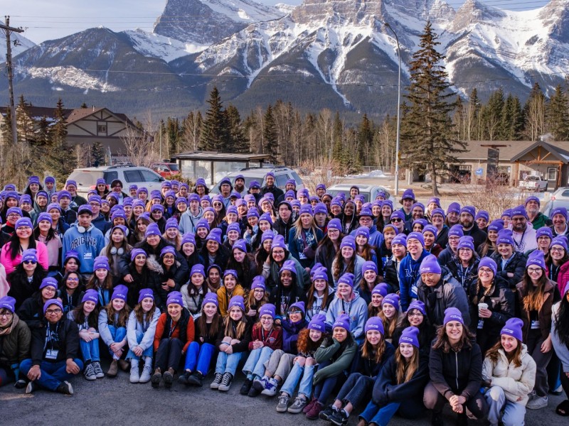 A large group of students and teachers gathered in a parking lot in front of a snowy mountain.
