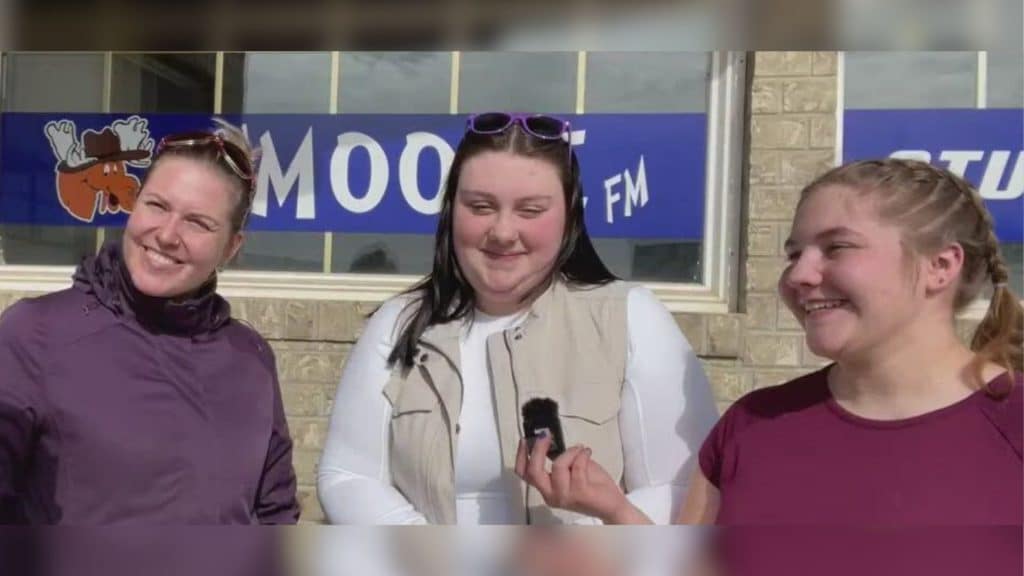 Three women standing in front of the Moose FM building, one of them holding a small microphone.