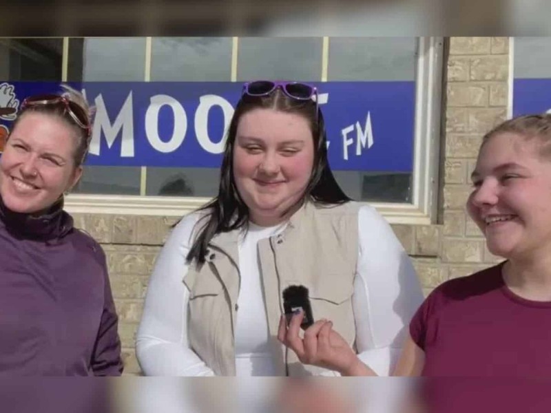 Three women standing in front of the Moose FM building, one of them holding a small microphone.