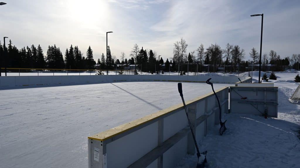 A picture of a boarded ice rink on a sunny day with snow shovels leaning against the boards.