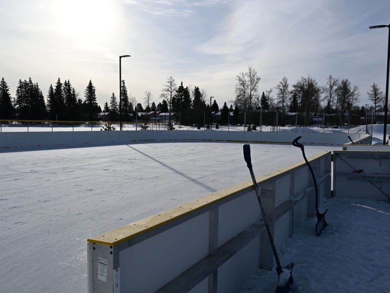 A picture of a boarded ice rink on a sunny day with snow shovels leaning against the boards.