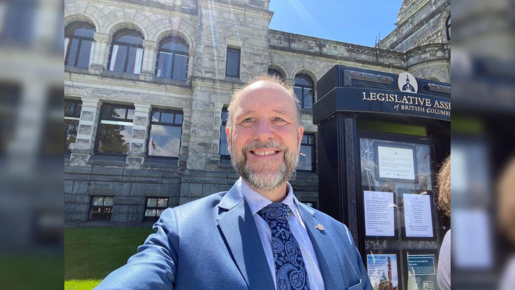 A late middle aged man in a blue suit with a grey beard.
