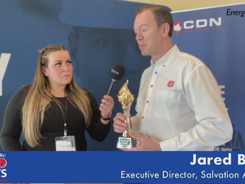 Two people standing in front of blue wall. Person on left holding a microphone up to person on right who is holding a trophy
